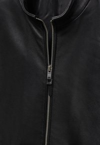 Primo piano di una giacca di pelle nera parzialmente aperta con una cerniera in metallo argentato visibile e fodera in tessuto nero.