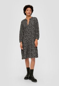 Schwarzes Blumenblattkleid mit langen Ärmeln, tiefem V-Ausschnitt, plissiertem Rock und strukturiertem Stoff. Getragen mit schwarzen Knöchelstiefeln.