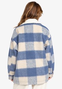 Eine blau-creamfarbene karierte Fleecejacke mit weicher Textur, die einen Kragen und knopfveredelte Manschetten aufweist.