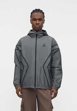 Mann trägt eine graue Zip-Hoodie-Jacke mit schwarzem Besatz und Logo, steht vor einem schlichten weißen Hintergrund.