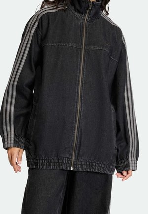 Personne portant une veste noire zippée avec trois bandes grises sur les manches et un pantalon noir assorti.