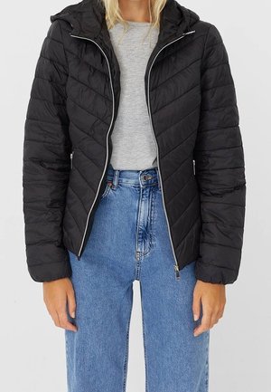 Kvinde iført en sort quiltet jakke med lynlås og hætte, grå t-shirt og højtaljede blå jeans mod en ensfarvet baggrund.