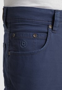 Jean en denim bleu foncé avec une texture lisse, présentant un design standard à cinq poches et un petit logo brodé sur la poche avant.