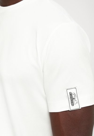 Vit kortärmad t-shirt på mörkhyad arm med en rektangulär tygetikett på ärmen som visar logotyp och text.