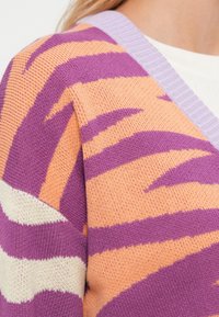 Πλεκτή ζακέτα με ρίγες σε μωβ, πορτοκαλί και κρεμ αποχρώσεις, με λαιμόκοψη σε λιλά ριμπ ύφανση και υφή στο ύφασμα.