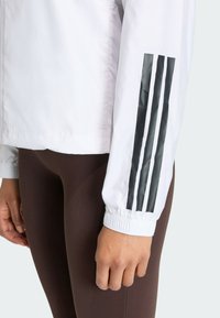White lightweight jacket with elastic cuffs, featuring three black stripes on the sleeve. Brown leggings with a smooth texture are visible.
