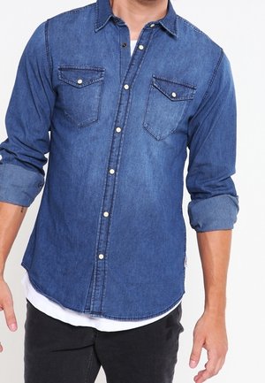 Homme portant une chemise en denim bleu à boutons avec des manches retroussées et un pantalon noir, se tenant contre un fond blanc uni.