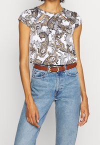 Mujer vistiendo una blusa de estampado paisley de manga corta metida dentro de unos pantalones vaqueros de tiro alto de color azul claro con un cinturón marrón y hebilla dorada.