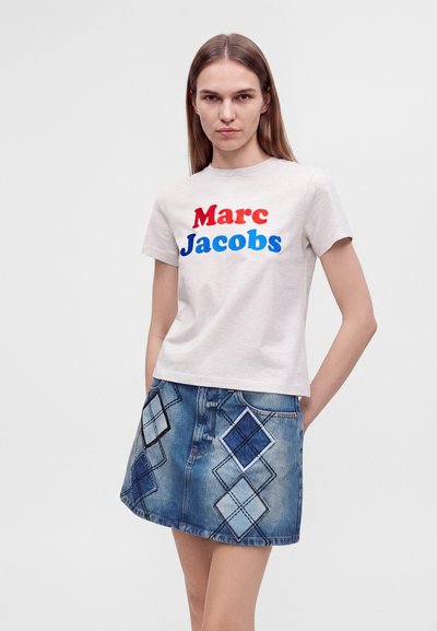Νέα γυναίκα με ίσια μαλλιά φοράει λευκό t-shirt Marc Jacobs με κόκκινα και μπλε γράμματα και μπλε τζιν φούστα με μπαλώματα σε σχήμα διαμαντιού.