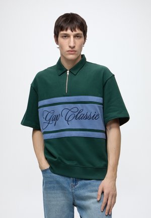 Jeune homme portant une chemise zippée manches courtes vert foncé avec le texte "Gap Classic" sur un panneau bleu, associée à un jean bleu clair.