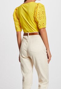 Gul bomullstopp med puffärmar som har ögonhålsblommönster, matchad med höga vita jeans och ett brunt bälte. Bakifrån visad.