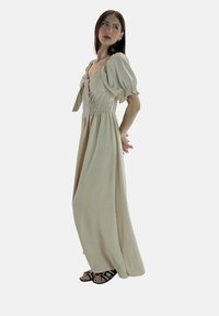 Lungo vestito beige con corpetto arricciato e maniche a sbuffo, presenta un laccio sul collo. Indossato con sandali neri con cinturini.
