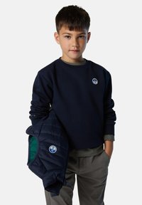 Felpa crewneck blu navy con una toppa rotonda, abbinata a un giubbotto imbottito blu navy con un interno verde e pantaloni grigi chiari.