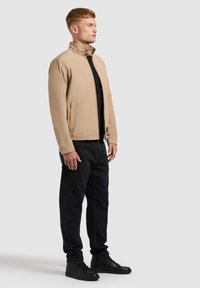 Moški stoji obrnjeno v desno, oblečen v bež jopico na zadrgo, črno majico, črne hlače in črne superge, na enobarvni svetli podlagi.