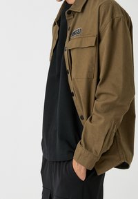 Giacca camicia in tela marrone con una tasca con un patch nero etichettato "EST 1988", indossata sopra una t-shirt nera e pantaloni neri.