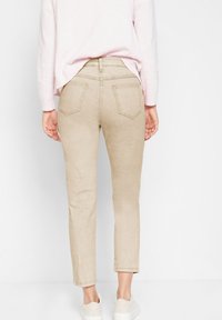 Žena v béžových slim-fit džínách, světle růžovém svetru a bílých teniskách stojící s uvolněnýma rukama po stranách, zády k divákovi.