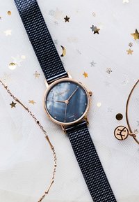 Rund armbåndsur med mørkeblå meshrem og mørkeblå urskive, guldfarvet urkasse, timeindikatorer af krystal, placeret på hvidt stof med guldstjerne- og måne konfetti.