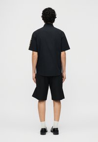 Άνδρας που φοράει μαύρο πουκάμισο με κοντά μανίκια και μαύρα σορτς μέχρι το γόνατο, στέκεται με την πλάτη προς τα εμάς σε απλό λευκό φόντο.