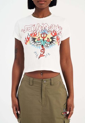 White cropped t-shirt featuring colorful graphic design with flames, dice, and a bat. Worn with olive green cargo-style skirt.