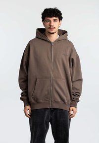 Felpa con zip di colore marrone, dotata di un ampio cappuccio, tasche a marsupio anteriori e polsini a coste. Realizzata in morbido tessuto con una vestibilità rilassata e oversize.