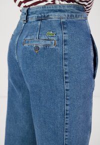 Jeans de mezclilla azul claro con una cintura alta, un bolsillo trasero con un logo de cocodrilo bordado en verde y un cierre de botón metálico negro.
