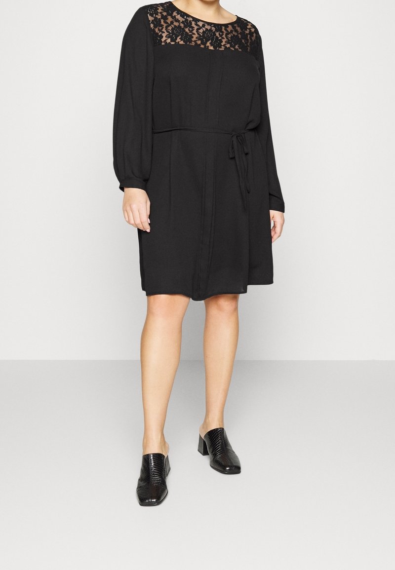 Svart klänning med spetsdetaljer vid axlarna, långa ärmar och knytning i midjan. Skorna är svarta, texturerade och har en låg blockklack. Enkelt, elegant design.