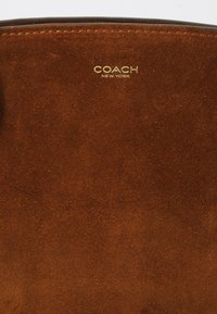 Borsa a tracolla in suede marrone con logo "COACH NEW YORK" in rilievo dorato. Texture liscia con design minimalista e cuciture curate.