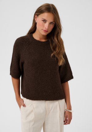 Femme aux longs cheveux ondulés portant un pull en laine marron foncé à manches aux coudes et un pantalon beige clair sur un fond uni.