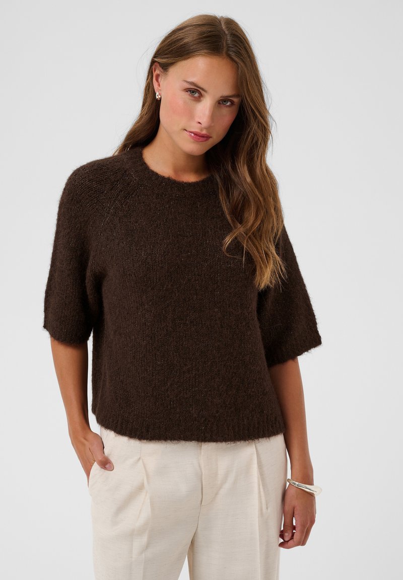 Femme aux longs cheveux ondulés portant un pull en laine marron foncé à manches aux coudes et un pantalon beige clair sur un fond uni.