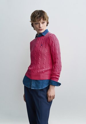 Person iført en lyserød kabelstrikket sweater over en blå denimknap-op-skjorte og marineblå bukser, stående mod en ensfarvet lys baggrund.