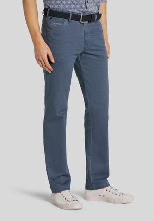 Mann trägt blaue gerade geschnittene Hose, einen dunkel gewebten Gürtel, weiße Sneakers und ein blau gemustertes Hemd vor einem neutralen Hintergrund.