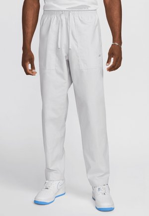 Mann trägt hellgraue Jogginghose mit Kordelzug und weiße Sneakers mit blauen Akzenten, steht vor einem einfarbigen hellen Hintergrund.