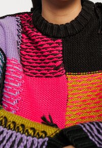 Multicolor gebreide trui met levendige roze, zwarte, gele, groene en paarse vlakken met textuurpatronen en een geribde halslijn.