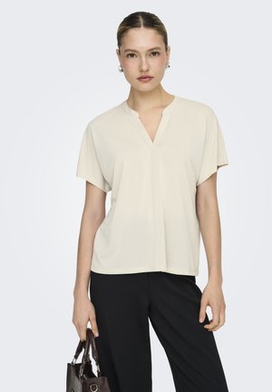 Mujer con blusa crema de manga corta y escote en V y pantalones negros, sosteniendo un bolso marrón oscuro texturizado con la mano izquierda.