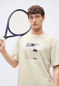 Beżowy bawełniany t-shirt z nadrukiem graficznym przedstawiającym abstrakcyjne kształty oraz słowo "BOSS", trzymany przez modela z rakietą tenisową.