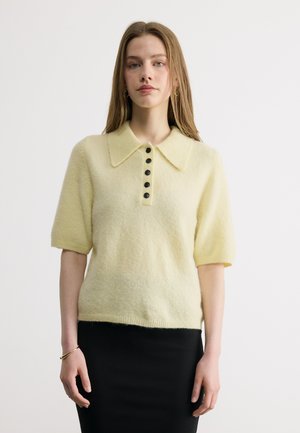 Νεαρή γυναίκα με μακριά μαλλιά, που φοράει ανοιχτό κίτρινο πουλόβερ με κοντά μανίκια και μαύρα κουμπιά, καθώς και μαύρη φούστα, μπροστά σε απλό φόντο.