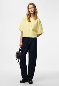 Gul ribbad kortärmad cardigan med knappar, kombinerad med mörka jeans med vida ben och en liten svart handväska med dragsko.