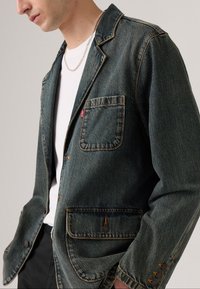 Hombre con chaqueta de mezclilla azul oscura con bolsillos delanteros, sobre una camisa blanca y pantalones negros, acompañado de un collar de cadena de plata.