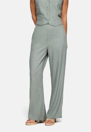 Mujer con pantalones anchos de pierna en verde claro y chaleco a juego abotonado, con sandalias de plataforma beige sobre un fondo blanco.