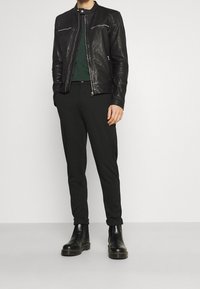 Černá kožená bunda se zipy, zelený top, úzké černé kalhoty a černé kotníkové boty. Oblečení se vyznačuje elegantními, minimalistickými designovými prvky.