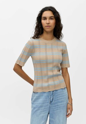 Kobieta o kręconych ciemnych włosach, ubrana w beżowy krótki sweter z niebieskimi paskami i jasnoniebieskie dżinsy, stojąca na jednolitym tle.