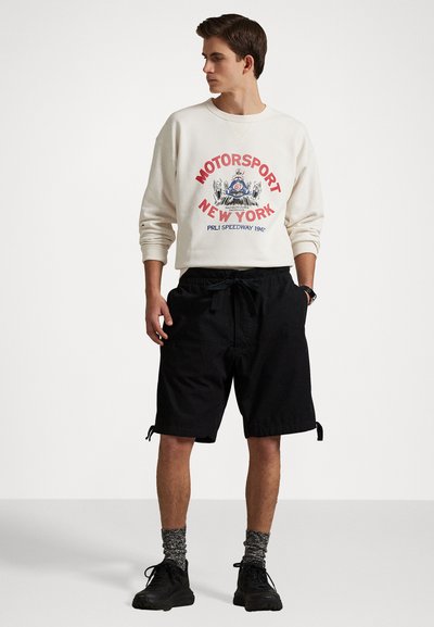 Witte sweatshirt met rode "MOTORSPORT NEW YORK" graphic, gecombineerd met zwarte shorts, textuur gebreide sokken en zwarte sneakers.