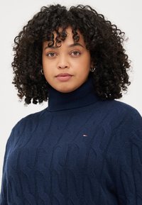 Eine junge Frau mit lockigem Haar (die einen dunkelblauen Rollkragenpullover trägt) blickt direkt in die Kamera vor einem schlichten Hintergrund.