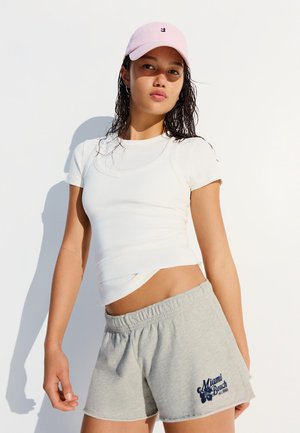 Ung kvinde iført en hvid tætsiddende t-shirt, grå Miami Beach-shorts og en lyserød baseballcap står mod en hvid baggrund.