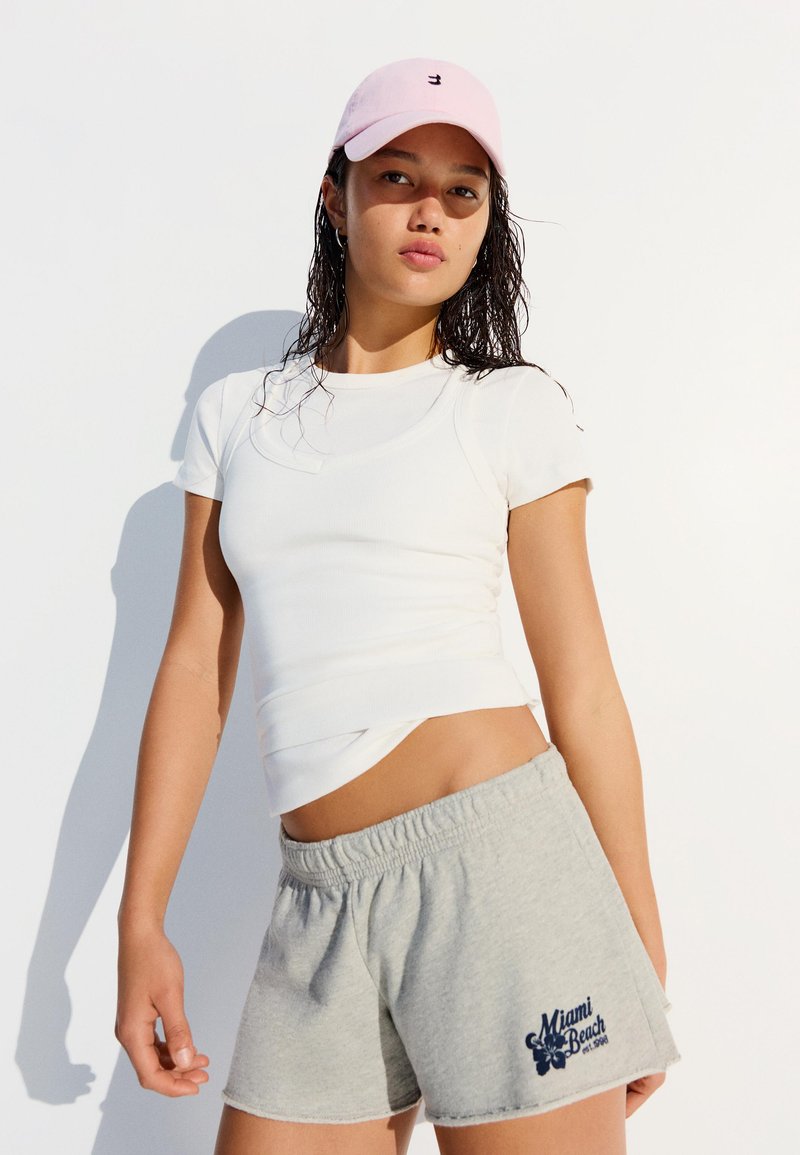 Jeune femme portant un t-shirt blanc ajusté, un short gris Miami Beach et une casquette de baseball rose clair, debout devant un fond blanc.