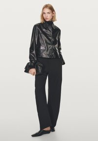 Zwarte leren jas met glanzende afwerking, grote kraag en knopen aan de voorkant. Gecombineerd met zwarte broek met wijde pijpen en een getextureerde clutch.