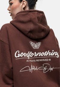 Felpa marrone realizzata in tessuto pesante. Presenta una grande scritta grafica bianca "Goodfornothing" e una farfalla sopra. Ha un cappuccio regolabile con coulisse.
