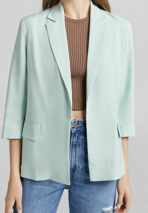 Frau trägt ein hellmintgrünes Blazer über einem braunen, gerippten Crop-Top und blauen, hoch taillierten, zerrissenen Jeans, steht vor einem schlichten Hintergrund.