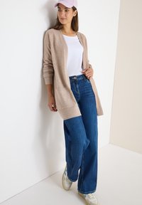 Svetle béžový dlouhý cardigan přes bílý tričko, v kombinaci s širokými modrými džíny a bílými teniskami se světlomodrými akcenty. Celkový vzhled doplňuje růžová čepice.