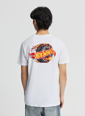 Persona che indossa una maglietta bianca con una ruota da corsa infuocata e il logo "Hot Wheels" in rosso e giallo sul retro, in piedi davanti a uno sfondo semplice.